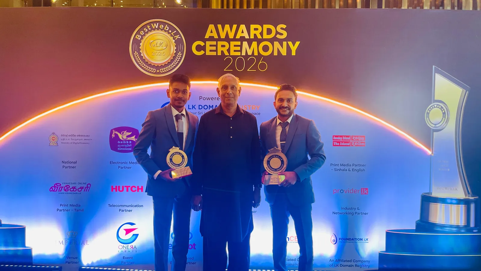 Two members of the Zonova team holding both trophies, with a guest, in front of the BestWeb.lk 2026 ceremony backdrop in Colombo.