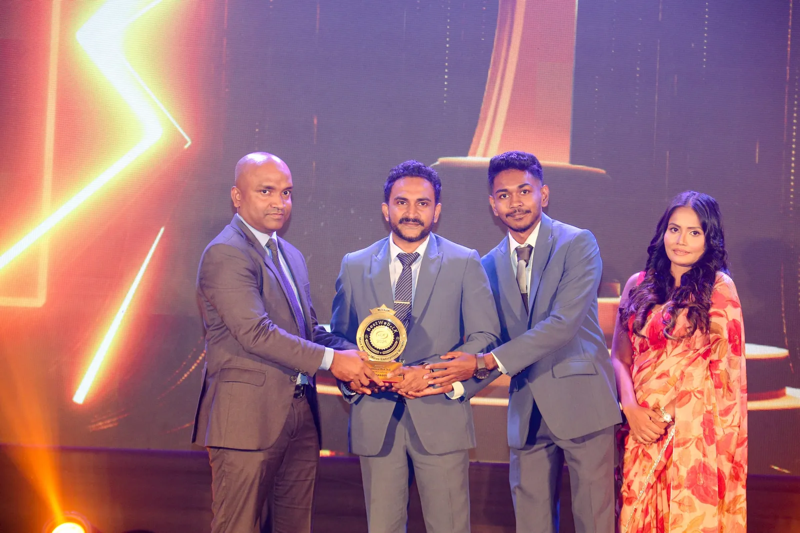 Three of the Zonova team and a BestWeb.lk presenter on stage, all of their hands together on the trophy.